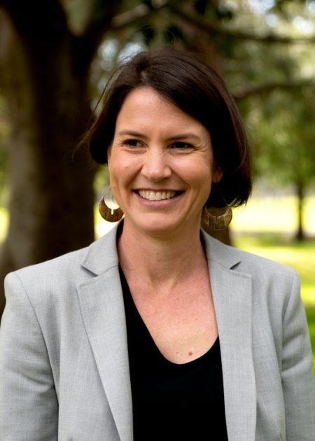 A woman with dark hair smiles while standing outdoors in a sunlit park. She is wearing large circular earrings, a light grey blazer, and a black top. Trees and green grass are visible in the blurred background.