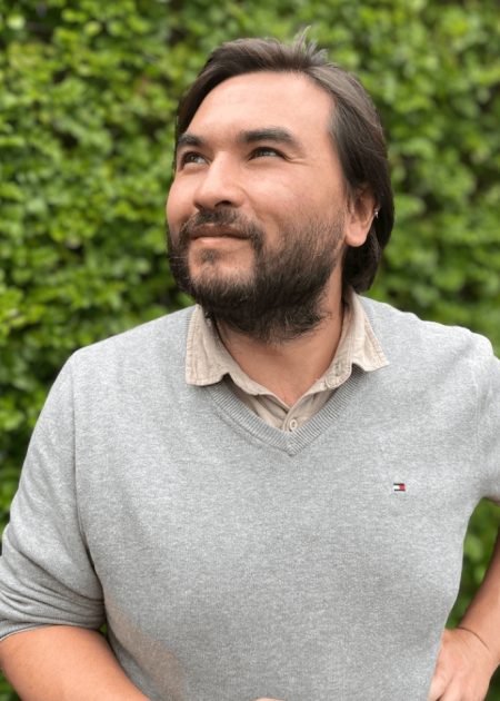 A man with dark hair and a beard looks upward with a thoughtful expression while standing in front of a lush green leafy wall. He is wearing a light grey sweater over a beige collared shirt, with one hand on his hip.