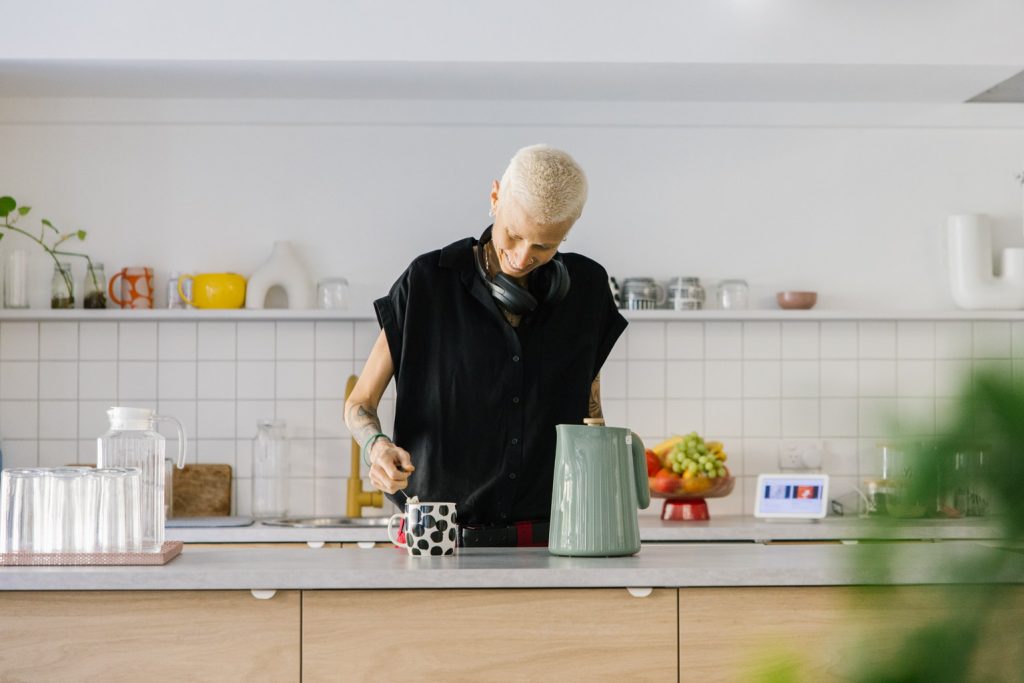 A person with short white hair wearing a black top, is in a kitchen making a cup of tea.