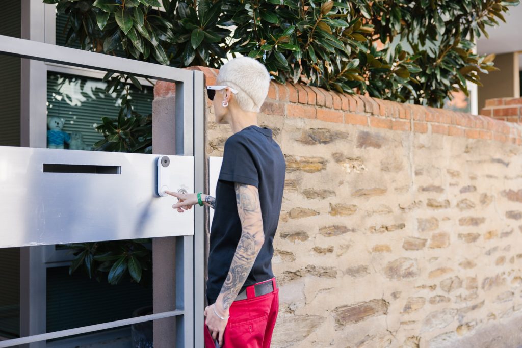 A person with short blonde hair wearing a black top and red pants is entering a front door of a home.