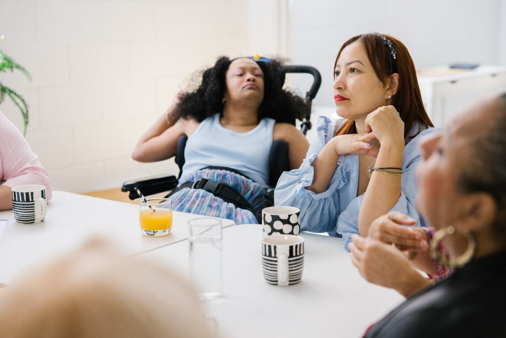 A young woman sits in a wheelchair next to another woman at a table