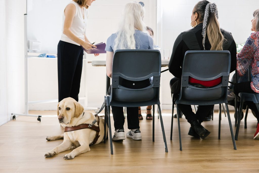 The backs of people sitting on chairs at a table. An assistance dog is lying on the floor.