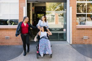 Three people are walking out of a building. One person is using a wheelchair.