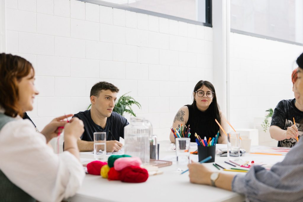 A group of people sitting at a table, talking to one another. There are craft supplies on the table.