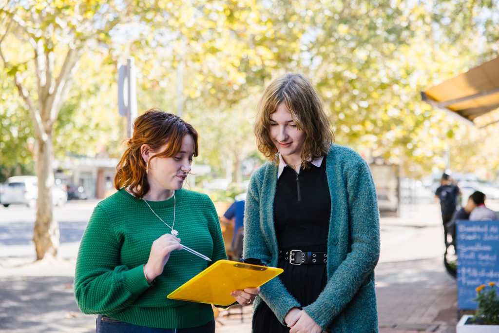 Two people are standing and talking in the street. One person is holding a clipboard, which the other person is looking at.