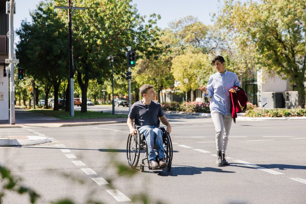 Two people are walking across the street, engaged in conversation. One is a man using a wheelchair.