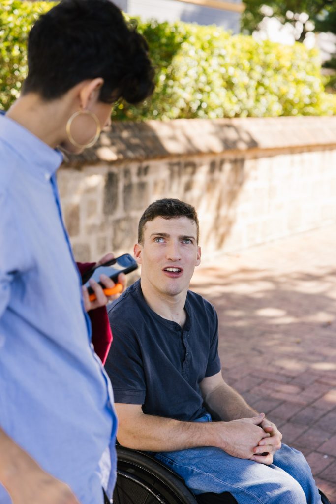 A man using a wheelchair is wearing a dark blue t-shirt ad jeans. He is talking to a person standing next to him, who has short back hair and a blue shirt.