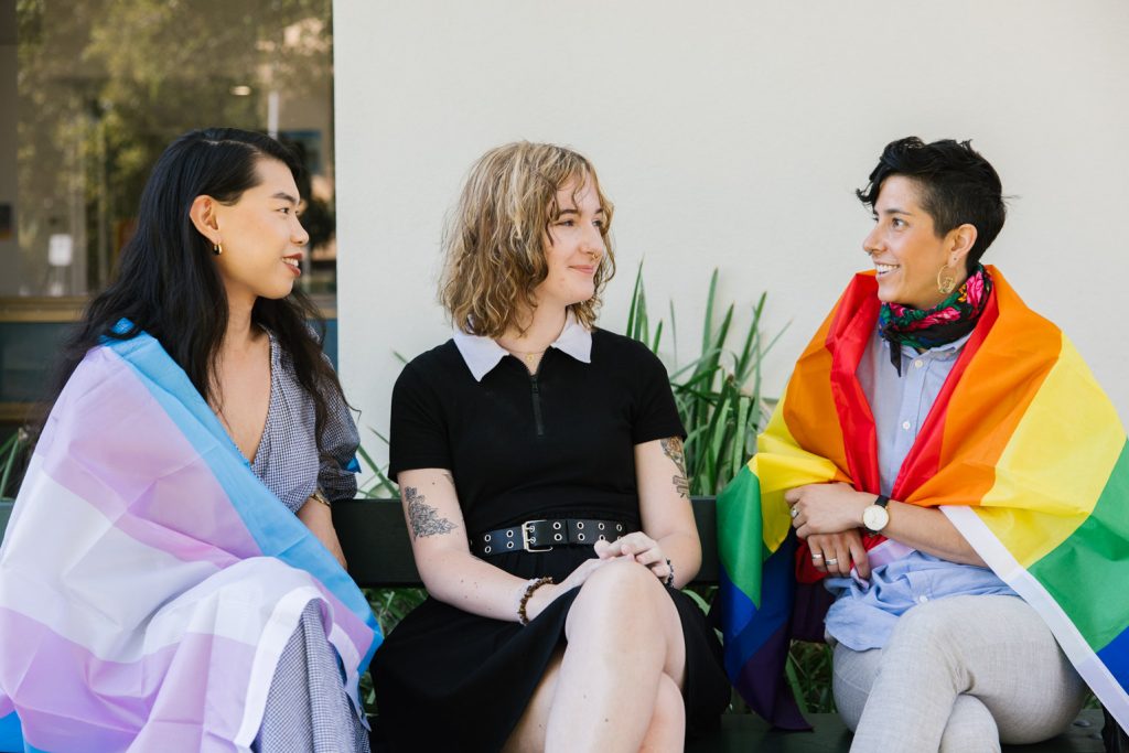 A group of three gender-diverse people are sitting on a bench talking. The person on the left has the transgender pride flag wrapped around their shoulders and the person on the right has the pride flag wrapped around their shoulders.