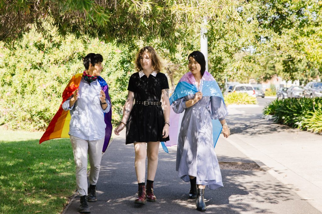 Three LGBTQIASB+ identifying people walking along a footpath wearing LGBTQIASB+ flags.