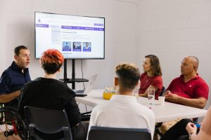 A group of people sit at a table and are looking at a screen.
