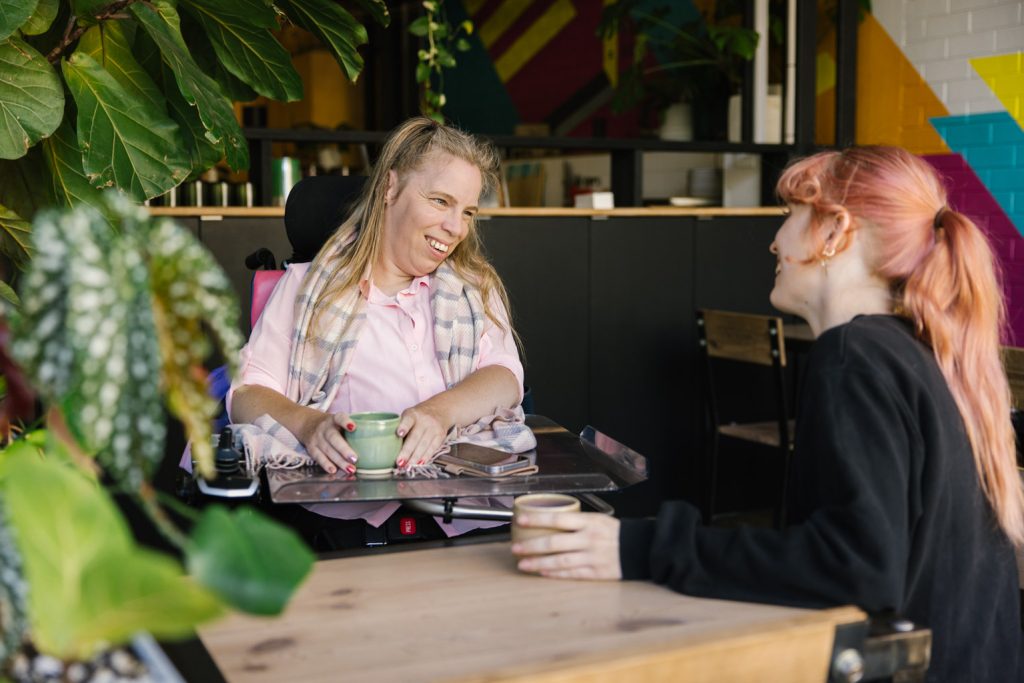 A woman using a wheelchair, with long blonde hair and a pink top talking to another woman with long orange hair and a black top. They are sitting at a cafe table.