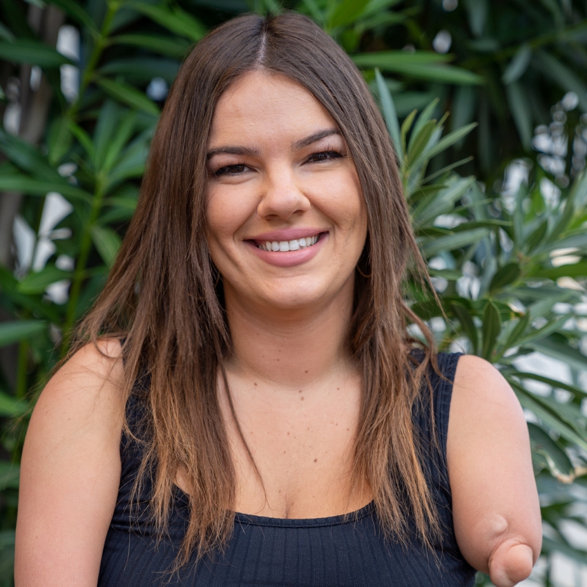 A woman with long brown hair and a black sleeveless top smiles at the camera, standing in front of green leafy plants. One of her arms is limb-different.