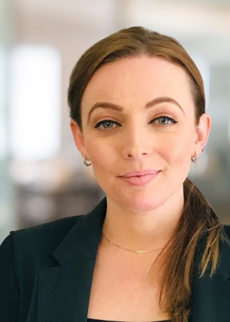 A woman with light brown hair tied in a low ponytail smiles softly at the camera. She is wearing a black blazer, small hoop earrings, and a thin gold necklace. The background is softly blurred, suggesting an office or professional indoor setting.