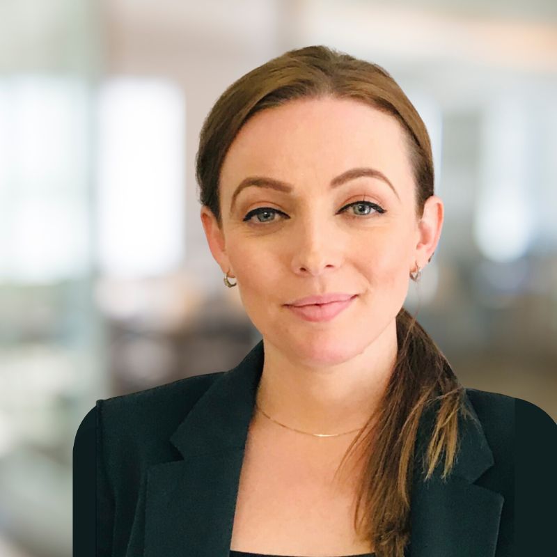 A woman with light brown hair tied in a low ponytail smiles softly at the camera. She is wearing a black blazer, small hoop earrings, and a thin gold necklace. The background is softly blurred, suggesting an office or professional indoor setting.