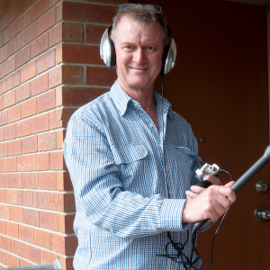 A man wearing large over-ear headphones stands outside near a brick wall, smiling at the camera. He is holding audio recording equipment, including a small recorder and a boom microphone. He wears a blue checkered shirt.