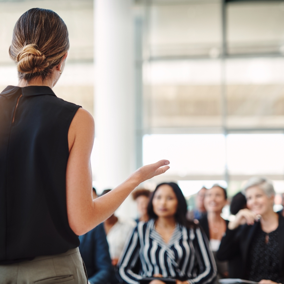A woman with her hair in a low bun stands with her back to the camera, speaking to a seated audience in a bright, modern meeting space. She gestures with one hand as the audience of professionals listens attentively. The setting includes large windows letting in natural light.