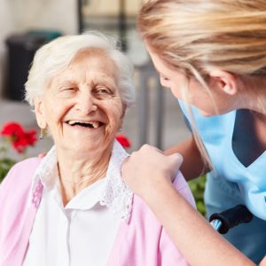 An elderly woman in a wheelchair is smiling and looking at a woman who is leaning over and looking back at her.