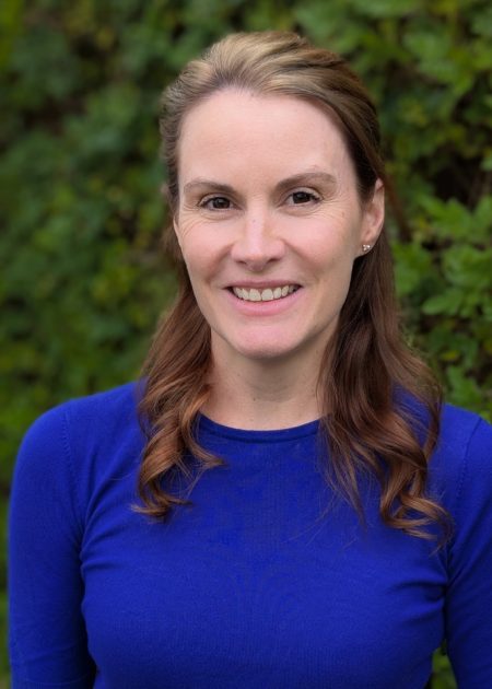 Smiling woman with long, wavy brown hair wearing a bright blue top, standing in front of a leafy green background.