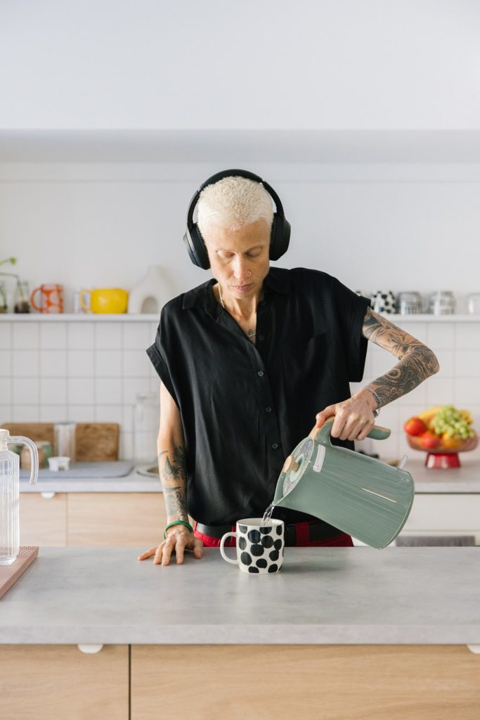 A person stands at a kitchen bench making a cup of tea. They have short bleached hair, tattoos on their arms, and wear a loose black top and headphones.