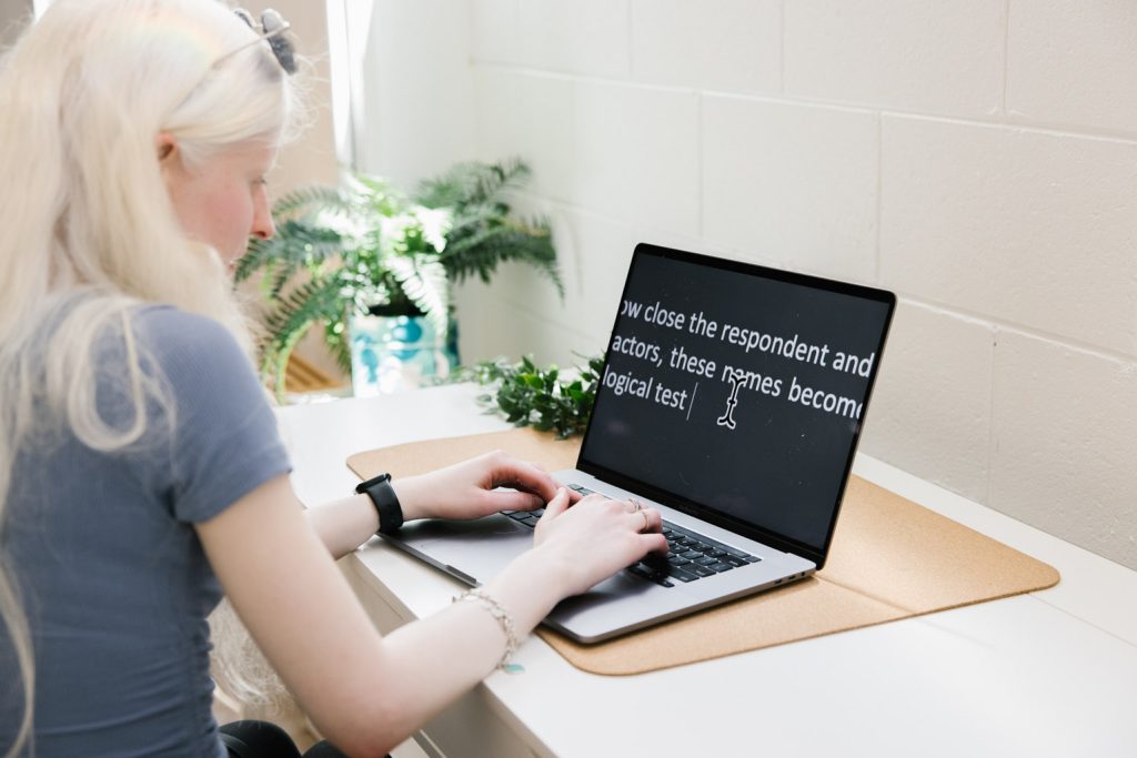 A woman with long, blonde hair types on a laptop at a white desk. The laptop screen displays enlarged white text on a black background, suggesting the use of assistive technology for low vision. The desk is decorated with green plants.