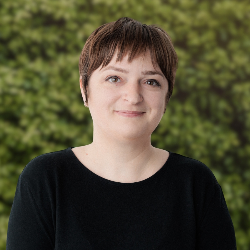A person with short brown hair and a gentle smile stands in front of a leafy green background. They are wearing a plain black top and looking directly at the camera.