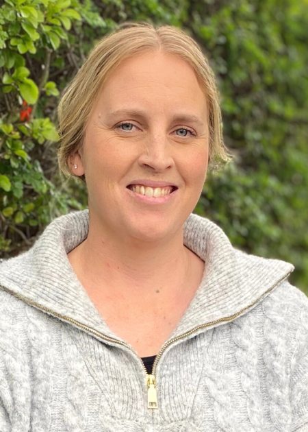 A blonde woman in a chunky light-grey cable-knit sweater smiles beside a leafy green hedge.