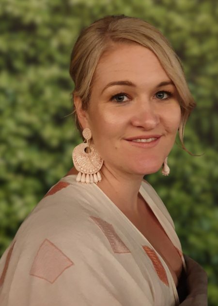 A woman with blonde hair styled to the side smiles softly at the camera. She wears large pale pink earrings and a light patterned top. The background is a softly blurred wall of green foliage.
