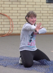 Child kneeling on the floor smiling and moving their hands, with a hula hoop behind them and fabric spread on the ground.