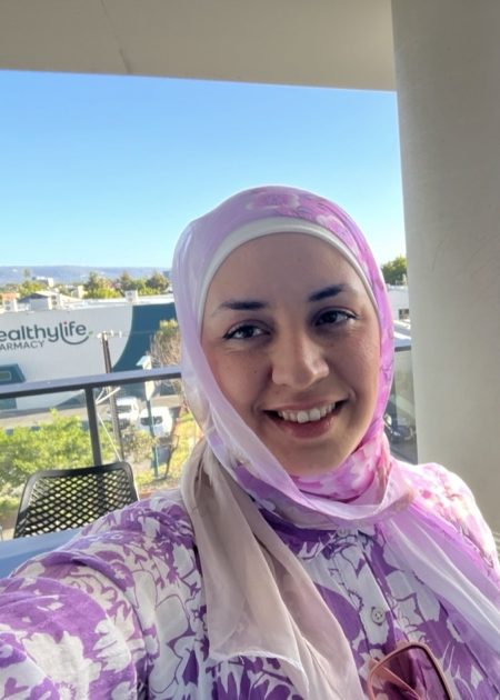 Salam smiles at the camera, wearing a pink and white floral hijab and matching patterned top, taking a selfie on a balcony with a sign, trees and hills visible in the background.
