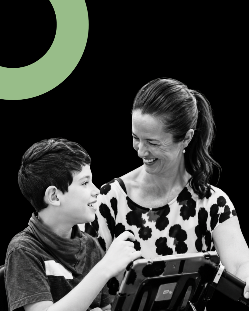 Black-and-white photograph of a smiling woman with dark hair in a ponytail looking at a young boy seated beside her. The boy smiles back while holding his hand near a communication tablet in front of them.
